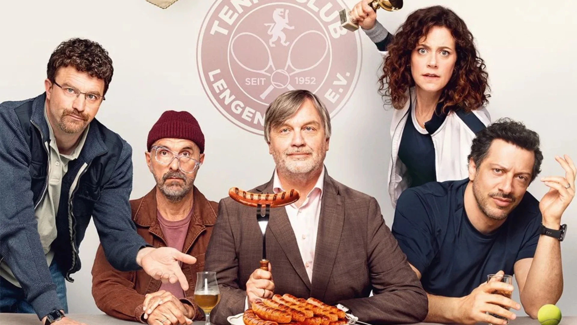Filmposter mit 5 Schauspielern die an einem Tisch sitzen und eine Wurst hochhalten